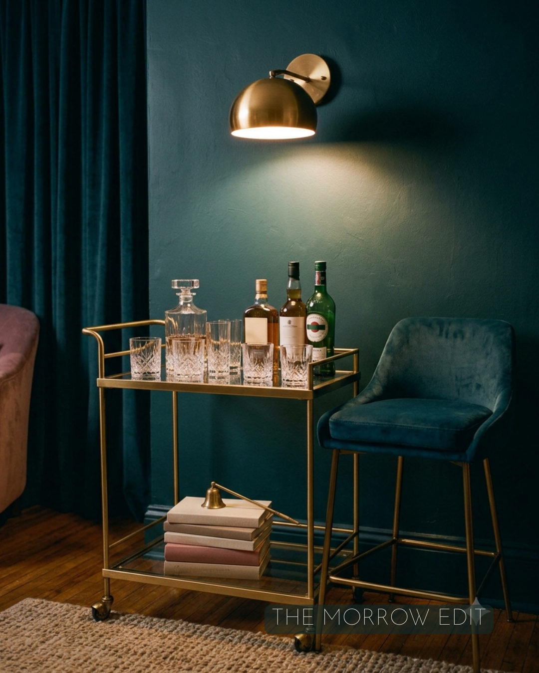 Moody brass bar cart against a deep teal wall, styled with a crystal decanter, glassware, and liquor bottles beneath a warm brass sconce, with a velvet bar stool alongside