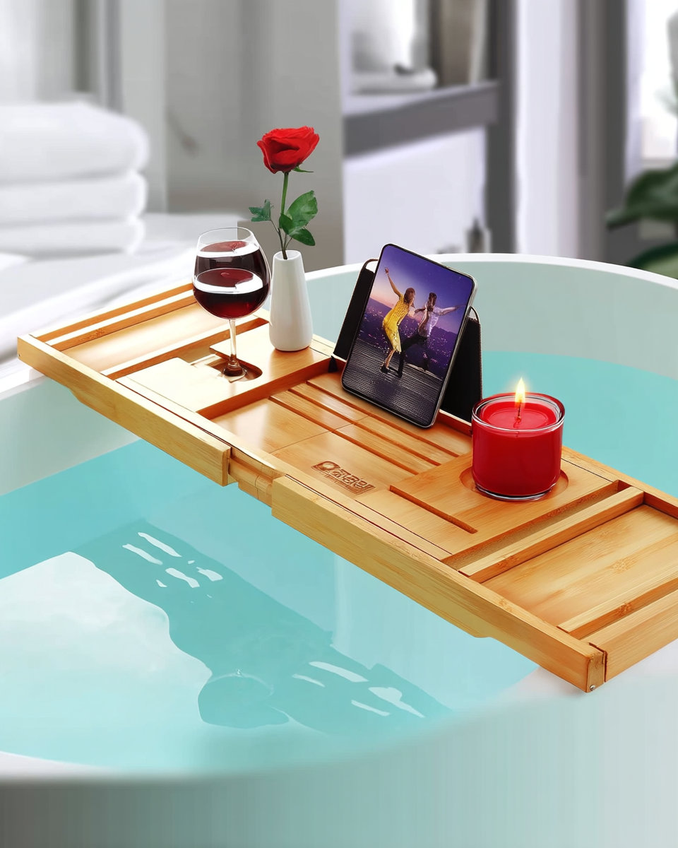Honey-toned bamboo bathtub caddy tray spanning a freestanding tub, holding a glass of wine, a tablet, and a lit candle over pale blue bathwater